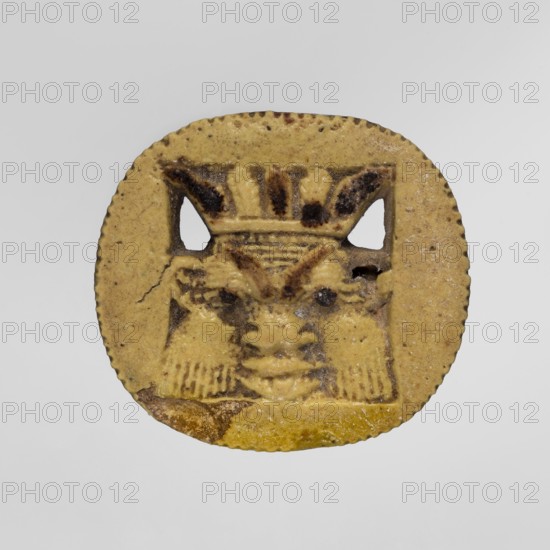 Amulet — plaque with the head of the god Bes. Amulet in the form of an oval, serrated, openwork plaque with a relief face of Bes wearing a feathered crown inside. Dark dots highlight the eyeballs and decorate some of the feathers., Egyptian amulet; Late Period, 664–332 BC. Type: amulet/plaque. Dimensions: height 4.3 cm, width 4.6 cm., A circular, flat object with a scalloped edge in shades of yellow and tan occupies the center of the image, showing a raised carved face motif with darker brownish accents highlighting facial features and decorative elements; the face has stylized eyes, a nose, and a mouth with carved lines suggesting hair or a headdress, and a few small holes or cutouts near the upper area; the surface shows some darker brown spots and slight chips along the lower edge against a light gray background.
