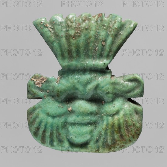 Amulet — plaque with the head of the god Bes;, Amulet in the form of a plaque with a relief depiction of the face of Bes, with a fan of feathers on the head and decorative curls of beard on both sides of the chin. Egyptian, Late Period, 664–332 BC. Materials: not stated. Function: amulet/pendant. Dimensions: height 3.8 cm, width 4.1 cm. Subjects: Bes myth., crowns, plaques, Egyptian deities., A carved green object with a weathered surface showing darker green and brownish spots, shaped like a stylized face with pronounced brow ridges, deep-set eye sockets, a rounded nose area, and a smiling mouth, topped by a fan-like crest with vertical grooves and flanked by broad, ribbed lower sections; the surface shows rough patches and small encrustations and the background is a neutral light gray.