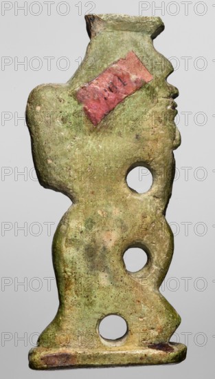 Amulet — the dancing god Bes; maker, Bes myth., Egyptian deities; feathers, dance, musical instruments; Egyptian religion; New Kingdom, ca. 1550–1069 BC; Egypt; amulet; height 6.7 cm, width 2.8 cm, A single vertical object shaped like a stylized profile of a human head and torso with a flat base, showing a greenish surface with patches of brown and beige discoloration, a torn piece of red material adhered near the upper area, three round holes arranged vertically through the object, a textured and slightly rough surface with spots of darker green and brown, and an overall weathered appearance.