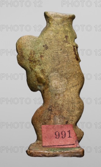 Amulet - dancing god Bes, Bes mythology. Figurine of Bes shown in right half-profile, head turned back; holds a tambourine in his left hand and grasps the base of his tail with his right. Four feathers on his head. Greenish faience. c. 1550–1069 BC; New Kingdom, Egypt. Amulet; height 4.6 cm, width 2.1 cm., A small, irregularly shaped object with a weathered, rough surface displayed against a plain light gray background; the object is primarily mottled brown with areas of greenish patina and some darker brown and reddish spots, showing uneven texture and worn edges; its silhouette has rounded protrusions and indentations, a narrow top widening into a bulbous middle and tapering toward a flat base; a small rectangular patch of muted pink is attached near the base.