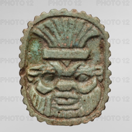 Amulet plaque with the head of the god Bes; plaque amulet, Egyptian deity Bes, wearing crowns. Late Period, Egypt, 664–332 BC. Height 3.4 cm; width 2.7 cm., A small oval object with a scalloped edge featuring a raised carved face with stylized facial features, including pronounced eyebrows, deep-set eyes, a broad nose, and a layered mouth area; the surface shows uneven texture with small pits and wear. The object displays a range of green tones from pale green to turquoise, with areas of brownish and reddish discoloration and darker green spots. The carving includes symmetrical vertical and curved ridges above the face that resemble a fan or headdress, and the relief varies in depth across the piece. The background behind the object is a smooth light gray.