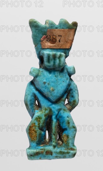 Amulet - god Bes, Bes myth., Figurine of Bes standing on slightly crooked legs, with his hands resting on his thighs, wearing a crown of four feathers. Blue faience, feather tips black. 664–332 BC; Late Period, Egypt. Amulet; height 4.8 cm, width 1.8 cm., A small carved figurine-like object with a predominantly turquoise blue color, showing areas of darker blue and greenish patina and some brownish discoloration, shaped with a crown-like top and a blocky head and torso, with protruding arm shapes and a skirted lower section that has a few vertical cutouts and rough textured spots, photographed against a plain light gray background.