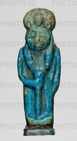 Amulet — figurine of a lion-headed goddess Sekhmet?, Sekhmet with a solar disk with a uraeus on her head, stands with a lotus scepter in her right hand left lowered. Egyptian religion; lionesses; Wajit myth.? / Sekhmet myth.?. Late Period, Egypt, 664–332 BC. Amulet; height 5.5 cm, width 1.8 cm., A small sculpted figure with a rounded head and a headdress, rendered in turquoise and green tones with patches of darker blue and brown weathering, shows a stylized face with closed eyes and a subtle nose. The figure has a pair of braided or tubular shapes hanging on either side of the head and down the front, and a raised element across the torso resembling an arm or symbol. The surface is rough and pitted in places, with areas of lighter greenish-blue and mottled darker green, and the base is a rectangular platform with bluish-green and brown discoloration.