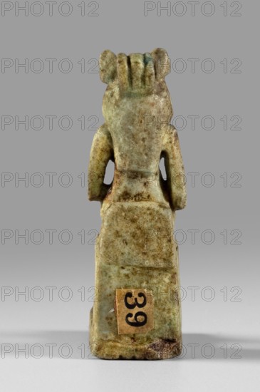 Amulet - figurine of a lion-headed goddess possibly Uajit or Sekhmet seated on a throne; Egyptian, Late Period, 664–332 BC. Material: amulet; dimensions: height 4.5 cm, width 1 cm., A small carved figurine shown from the back, with a weathered surface in shades of green, pale green, beige, brown, and hints of bluish-green; the figure has a rectangular base and a tapered torso with stylized arms that curve down along the sides, a head with short vertical ridges at the top and rounded ear-like shapes, and surface mottling and spots in darker brown and tan; a small rectangular patch of tan material with dark markings is affixed near the lower back of the base; the background is a smooth mid-gray that fades slightly toward the bottom.