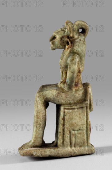 Amulet - figurine of a lion-headed goddess sitting on a throne Uajit?, lionesses, Egyptian deities, Uajit myth., thrones, Sekhmet myth., 664–332 BC; Late Period, Egypt, amulet, height 4.5 cm, width 1 cm, A small sculptural figure of a seated animal-headed person is shown in profile on a neutral gray background, rendered in a weathered greenish-beige material with areas of brownish discoloration and darker green speckling; the figure sits on a rectangular stool with a flat base, the legs extended forward and the hands resting on the thighs, and the head has a curled element at the back with an opening through it, while the surface shows a slightly rough, aged texture and subtle shadows along the contours.