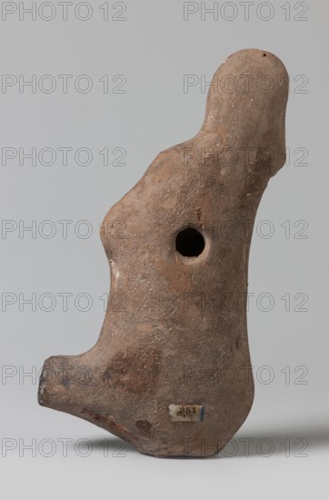 Lamp with a depiction of Athena-Neith, Mithra mythology, Attys mythology, Phrygian caps iconography, figural lamp. Lamp decorated with a youth, standing on its container, leaning against a column topped with a bird's head bent over a palm capital. The young man stands cross-legged, dressed in a garment that can be interpreted as a chiton or as Roman armor with pteryges, he has a Phrygian cap, on his head, and his right hand rests on a circular object a tambourine or rather a shield. 1st–3rd century, Egypt, oil lamp; height 19.5 cm, width 9.7 cm, depth 4 cm., A weathered, irregularly shaped object made of rough, brown material stands upright against a plain light gray background; the object has an overall earthy brown color with patches of darker brown and small areas of lighter, almost tan tones, a rounded top that tapers into a bulging midsection with a circular hole, a protruding lower extension that curves outward, a small rectangular label attached near the base with a strip of blu...