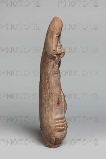 Lamp with a depiction of Athena–Neith, Lamp decorated with a youth, standing on its container, leaning against a column topped with a bird's head bent over a palm capital. The young man stands cross-legged, dressed in a garment that can be interpreted as a chiton or as Roman armor with pteryges, wearing a Phrygian cap, and resting his right hand on a circular object a tambourine or shield. Mithra mythology, Attys mythology, Phrygian caps iconography, figural lamp, Class.Gal.Scen., Athena mythology. 1st–3rd century, Egypt, oil lamp; height 19.5 cm, width 9.7 cm, depth 4 cm, A tall, narrow sculpture photographed against a plain light gray background, rendered in earthy brown and beige tones with some darker brown spots; the surface appears textured and weathered with visible rough patches and small holes; the form tapers toward the top and widens toward the base, with subtle carved contours suggesting a rounded top, a recessed midsection, and a series of horizontal ridges near the lower ...