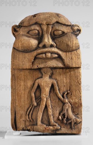 Magical stele topped with the mask of the god Bes. Magical stele cippus depicting in the lower register a naked Horus-Child in a scene of triumph over dangerous creatures. In his right hand, Horus likely holds two snakes, in his left a four-legged animal—perhaps a hyena or a lion. Horus stands on a crocodile. The depiction is related to the myth of a venomous snake attacking the infant Horus during the absence of his mother, Isis; the initiator of the attack was Seth, brother of Osiris, whom young Horus fought to avenge his father's death. The upper part of the monument depicts the face of the god Bes. 664–30 BC; Late Period–Ptolemaic Period, Egypt. Stele; height 20.5 cm, width 12.5 cm., A carved wooden plaque showing a large stylized face at the top with pronounced cheekbones, a broad nose, full lips, and deep-set eye shapes, rendered with visible carving marks and darkened recessed lines; below the face a carved human figure stands with an elongated torso and limbs, one arm extended ...
