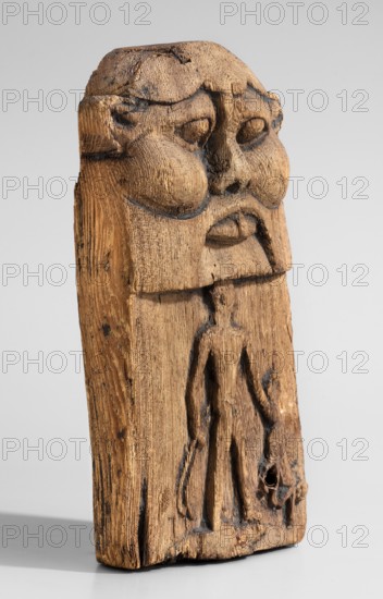 Magical stele topped with the mask of the god Bes. Magical stele cippus depicting in the lower register a naked Horus-Child in a scene of triumph over dangerous creatures. In his right hand, Horus likely holds two snakes; in his left a four-legged animal—perhaps a hyena or a lion. Horus stands on a crocodile. The depiction relates to the myth of a venomous snake attacking the infant Horus during the absence of his mother Isis; the initiator of the attack was Seth, brother of Osiris, whom young Horus fought to avenge his father's death. The upper part of the monument depicts the face of the god Bes. Date: 664–30 BC Late Period–Ptolemaic Period. Material/object type: stele. Dimensions: height 20.5 cm; width 12.5 cm., A carved wooden figure shown upright against a pale gray background, with visible grain and textured tool marks; the top portion features a stylized face with pronounced rounded cheeks, deep-set eye hollows, a broad nose, and a downturned mouth, all carved in relief and show...