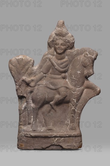 Figurine of Harpocrates-Heron on horseback, figurine of a deity sitting on a horse with a wreath and a double crown on his head, dressed in a short military tunic and cloak. The horse is walking to the right, the deity is facing the viewer, to the right. Behind the horse's rump is a palm tree, 2nd century, Egypt, height 29 cm, width 19.3 cm, Egyptian deities, Harpocrates mythology, Heron mythology, horses., A weathered stone relief in shades of brown and gray shows a carved figure seated on an animal resembling a horse. The figure has a rounded face and stylized hair or headdress and wears draped garments suggested by carved folds across the torso. The animal has a defined head with a visible ear and snout and a molded mane. The relief surface displays areas of lighter discoloration and small cracks, with rough, chipped edges along the top and sides. The base is rectangular and slightly raised, with worn contours and patches of lighter mineral streaking. The background behind the carvi...