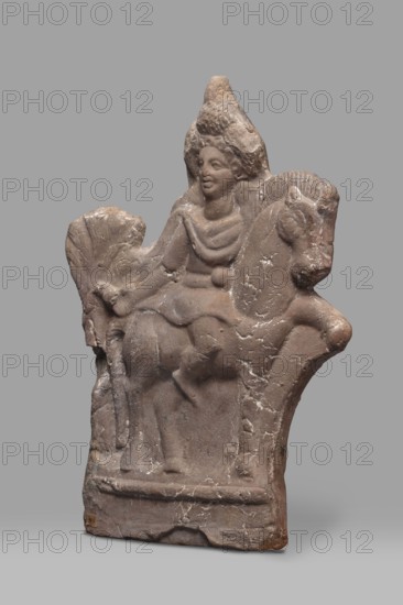 Figurine of Harpocrates-Heron on horseback, Egyptian deities, Harpocrates mythology, Heron mythology, horses. Figurine of a deity sitting on a horse with a wreath and a double crown on his head, dressed in a short military tunic and cloak. The horse is walking to the right, the deity is facing the viewer, to the right. Behind the horse's rump is a palm tree, 2nd century, Egypt, figurine, height 29 cm, width 19.3 cm, The image shows a carved stone relief of a figure seated on an animal, set against a plain gray background; the stone has a weathered, rough texture with visible cracks and chips, and its color ranges across shades of brown and tan with lighter beige highlights and small areas of white veining; the seated figure wears a draped garment and a tall headdress and has a rounded face with softly rendered features; the animal beneath the figure has a rounded body and a head with a pronounced snout and ears; the base of the relief is rectangular with an angled edge and shows surfac...