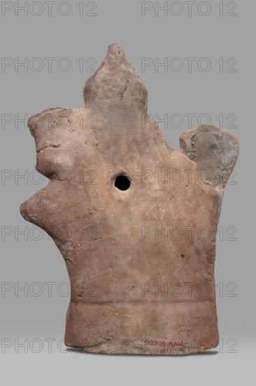 Figurine of Harpocrates-Heron on horseback, Egyptian deity Harpocrates combined with Heron, deity seated on a horse wearing a wreath and a double crown, dressed in a short military tunic and cloak. The horse walks to the right while the deity faces the viewer to the right; a palm tree appears behind the horse's rump. 2nd century, Egypt; terracotta figurine, height 29 cm, width 19.3 cm., A weathered clay object with an irregular, roughly vertical silhouette and an overall earthy brown color with patches of gray and lighter beige, showing a rough, pitted surface texture and uneven edges; a rounded protrusion on one side and another irregular lobed extension near the top, plus a central circular hole; the base is broader and slightly darker with subtle surface cracks and a few small darker spots; the background is a flat gray.