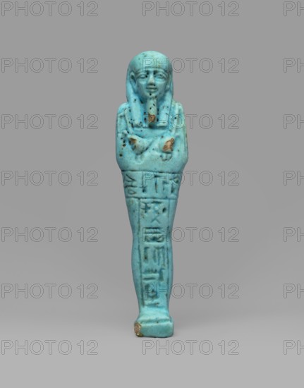 Ushabti Wahibre-em-achet. Mummy-shaped funerary figurine shabti made of blue faience with black details. Stands on a base, supported by a pillar at the back. Arms crossed on the chest, right folded over left; from under the shroud only clenched hands are visible. Right hand holds a hoe, left hand a pickaxe; a basket is slung over the left shoulder. Engraved tools. Smooth three-part wig with longer front strands; straight braided beard. Large eyes and a slightly smiling mouth. Engraved T-shaped inscription: one line running around the figure at hip level and one column on the front of the legs reading, Shd Osiris the father of God, priest ... Uah-ib-Re-em-achet born of Ta-net-Amun. Ca. 664–525 BC; Late Period, 26th Dynasty, Egypt. Height 13.4 cm; width 3 cm., A small carved figurine of a standing human figure shown full-length, rendered in a bright turquoise blue with areas of darker blue-green and small brown spots and chips; the figure has a stylized headdress and a defined face with ...