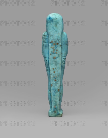 Ushabti Wahibre-em-achet. Mummy-shaped funerary figurine shabti made of blue faience with black details. Stands on a base, supported by a back pillar. Arms crossed on the chest, right folded over left; hands clenched into fists visible from under the shroud. Holds a hoe in the right hand and a pickaxe in the left; a basket is slung over the left shoulder. Engraved tools. Wears a smooth, three-part wig with longer front strands; straight braided beard. Large eyes and a slight smile. Engraved inscription arranged in a T-shape — a line around the figure at hip level and a column on the front of the legs: Shd Osiris the father of God, priest ... Uah-ib-Re-em-achet born of Ta-net-Amun. Circa 664–525 BC; Late Period, 26th Dynasty, Egypt. Height 13.4 cm; width 3 cm., A vertical figurine-shaped object standing upright against a plain background, predominantly turquoise blue with areas of darker blue-green and small brownish spots; the object has a rounded top, narrow shoulders, elongated body ...