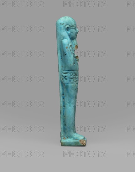 Ushabti Wahibre-em-achet, Back pillar, hieroglyphs, Wahibre-em-achet ca. 664–525 BC. Mummy-shaped funerary figurine shabti made of blue faience with black details. Stands on a base, supported by a pillar at the back. Arms crossed on the chest, right folded over left; from under the shroud only the hands clenched into fists are visible. Right hand holds a hoe, left holds a pickaxe; a basket is slung over the left shoulder. Engraved tools. Smooth three-part wig with front strands longer than the back; straight braided beard. Large eyes and a prominent mouth stretched in a slight smile. Engraved inscription arranged in a T-shape—one line around the figure at hip level and one column on the front of the legs—reading: Shd Osiris the father of God, priest ... Uah-ib-Re-em-achet born of Ta-net-Amun. 664–525 BC; Late Period, 26th Dynasty, Egypt. Height 13.4 cm, width 3 cm., A slender standing figurine viewed in profile against a neutral gray background, rendered in a bright turquoise-blue with...