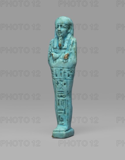 Ushabti Wahibre-em-achet. Mummy-shaped funerary figurine shabti, ca. 664–525 BC, Late Period, 26th Dynasty, Egypt. Made of blue faience with black details. Stands on a base, supported by a pillar at the back. Arms crossed on the chest, right folded over left. From under the shroud only the hands clenched into fists are visible; right hand holds a hoe, left a pickaxe; a basket is slung over the left shoulder. Engraved tools. Smooth three-part wig with longer front strands than the back. Straight, braided beard. Large eyes and a mouth stretched in a slight smile. Engraved inscription arranged in a T-shape — one line running around the figure at hip level and one column on the front of the legs: Shd Osiris the father of God, priest ... Uah-ib-Re-em-achet born of Ta-net-Amun. Height 13.4 cm; width 3 cm., A small turquoise-blue figurine shaped like a standing mummiform person with a rounded head and stylized facial features, crossed arms, and a long narrow body; the surface shows some darke...