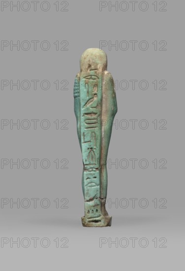 Ushabti Psamtyk-seneb, wigs, funerary figurine, hieroglyphs, beards facial hair, Psamtyk-seneb ca. 747–332 BC, back pillar/pilaster, grave goods, mummy-shaped figurine standing on a base, back supported by a pillar. Arms crossed on the chest, right folded over left. In the right hand a long-bladed hoe, in the left a pickaxe; handles of both hoes rolled up at the ends. A bag slung over the left shoulder on a single string on the back. Three-part smooth wig; long tapering beard braided into a plait. Engraved inscription in one column on a pilaster reading Osiris Psammetik-seneb born of Jerou justified by the voice. 664–332 BC; Late Period, Egypt. Shabti; height 12.5 cm, width 3 cm., A small upright figurine viewed from the back, with a slender elongated body and a rounded head, standing on a short rectangular base; the surface shows a weathered, slightly rough texture with visible chips and small cracks; the left side of the figure has a series of incised vertical shapes and shallow reli...