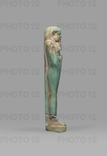 Ushabti Psamtyk-seneb, wigs, funerary figurines, hieroglyphs, beards facial hair, Psamtyk-seneb ca. 747–332 BC, back pillars, grave goods, mummy-shaped figurine standing on a base, back supported by a pillar/pilaster. Arms crossed on the chest, the right folded over the left. In the right hand is a long-bladed hoe, in the left a pickaxe. The handles of both hoes are rolled up at the ends. On the back is a bag, slung over the left shoulder on a single string. On the head is a three-part, smooth wig. The beard is long, tapering toward the bottom, and braided. Engraved inscription in one column on a pilaster reading Osiris Psammetik-seneb born of Jerou justified by the voice. 664–332 BC; Late Period, Egypt; shabti; height 12.5 cm, width 3 cm., A standing figurine seen in profile against a plain gray background, the figure has a long, narrow form with a headdress and arms folded across the chest, the surface shows a patina with shades of green and turquoise, along with areas of brown and b...