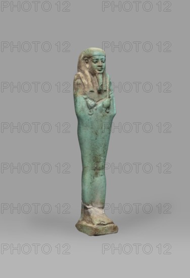Ushabti Psamtyk-seneb, Mummy-shaped funerary figurine shabti, standing on a base, back supported by a pillar/pilaster. Arms crossed on the chest right folded over left. Right hand holds a long-bladed hoe, left hand a pickaxe; handles rolled up at the ends. A bag is slung over the left shoulder on a single string. Wears a three-part smooth wig; long tapering beard braided into a plait. Engraved inscription in one column on the pilaster: Osiris Psammetik-seneb born of Jerou justified by the voice. Late Period, Egypt ca. 664–332 BC. Height 12.5 cm, width 3 cm., A small standing figurine with a teal-green surface and areas of brown and beige patina, showing a human figure with a headdress and crossed arms, a rounded head with facial features, a tapered body mounted on a rough base, set against a plain gray background.
