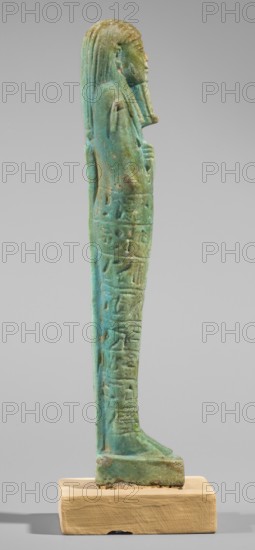 Ushabti of Semen-Ptah-Psamtyk. Wigs, funerary figurine mummy-shaped, hieroglyphs, beards facial hair, hoes, back pillar/pilaster. Semen-Ptah-Psamtyk ca. 747–332 BC. Book of the Dead motifs; grave goods. Standing on a base, supported at the back by a pillar. Hands crossed on the chest, right folded over left. In the right hand a hoe with a narrow blade; in the left hand a pickaxe. A basket slung on a single string over the left shoulder. Three-part wig styled in narrow strands ending in a horizontal hairline at the front. Chin tapers downward with braided curl; almond-shaped eyes, broad nose, full slightly pouty lips. Engraved inscription in eight lines around the figurine text begins: O you shabti! If Osiris Semen-Ptah-Psammetik, born of Har-pe-khrad-ankhet... to cultivate the field, to irrigate the bank, to transport sand from west to east and from east to west. Here I am, you will say.. 8 lines of text. 664–332 BC, Late Period, Egypt. Shabti. Height 19 cm; width 4.9 cm., A vertical s...