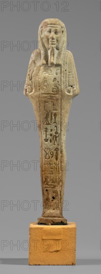 Hornakht's shabti, back pillars, hieroglyphs, wigs, beards facial hair. Hornakht ca. 525–30 BC. Funerary figurine mummy-shaped shabti, standing on a base, back supported by a pillar/pilaster. Arms crossed on the chest, right folded over the left. In the right hand a long-bladed hoe and two strings from which hangs a semicircular basket on the back, slightly above the elbow; in the left hand a whip. Hands and tools engraved. Long, three-part smooth wig; long straight beard of uniform width. Face with coarse features, roughly defined eyes, wide nose, prominent mouth set in a slight smile. Right ear significantly larger than the left. Inscription carved in a single column on the front from chin to feet, naming the deceased Hornakht and his mother content otherwise unclear. Date: 664–30 BC Late Period–Ptolemaic. Egypt. Shabti. Height 11.6 cm; width 3.1 cm., A standing statuette of a human figure with a headdress and stylized beard, rendered in a weathered beige and light brown stone with d...