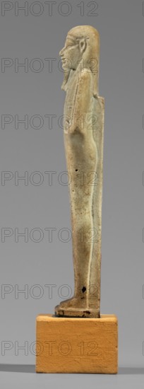 Hornakht's shabti, back pillars, hieroglyphs, wigs, beards facial hair; Hornakht ca. 525–530 BC; funerary figurine, mummy-shaped, standing on a base, back supported by a pillar/pilaster. Arms crossed on the chest, right folded over the left. Right hand holds a long-bladed hoe and two strings from which hangs a semicircular basket on the back, slightly above the elbow; left hand holds a whip. Hands and tools are engraved. Long, three-part smooth wig; long straight beard of uniform width. Face with coarse features, roughly defined eyes, wide nose, prominent mouth with a slight smile. Right ear significantly larger than the left. Inscription carved in a single column on the front from chin to feet, naming the deceased Hornakht and his mother content otherwise unclear. Date: 664–30 BC Late Period–Ptolemaic Period, Egypt; shabti; height 11.6 cm, width 3.1 cm., A slender vertical statuette in muted beige and pale green tones stands on a rectangular ochre base, showing a side profile of a hum...