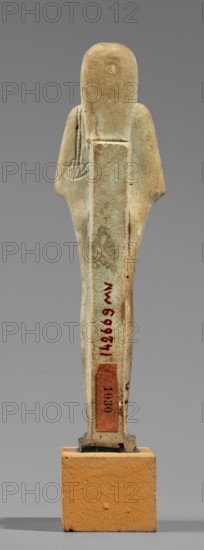 Hornakht's shabti, Back pillars, hieroglyphs, wigs, beards facial hair. Hornakht ca. 525–30 BC. Funerary figurine mummy-shaped shabti, standing on a base, back supported by a pillar/pilaster. Arms crossed on the chest, right folded over the left. In the right hand a long-bladed hoe and two strings supporting a semicircular basket on the back, slightly above the elbow; in the left hand a whip. Hands and tools are engraved. Head wears a long, three-part smooth wig. Beard long, straight, uniform width. Face with coarse features: roughly defined eyes, wide nose, prominent mouth with a slight smile. Right ear significantly larger than the left. Inscription carved in a single column on the front, extending from chin to feet; content unclear, names the deceased Hornakht and his mother. Date: 664–30 BC Late Period–Ptolemaic Period, Egypt. Type: shabti. Height 11.6 cm; width 3.1 cm., A small upright figurine stands on a rectangular base, shown from the back against a neutral gray background. Th...