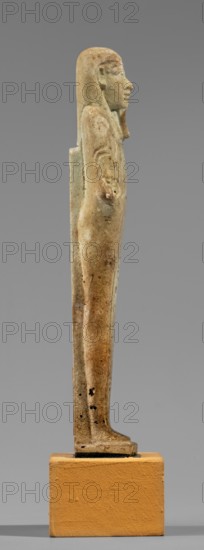 Hornakht's shabti, back pillars, hieroglyphs, wigs, beards facial hair. Hornakht ca. 525–30 BC. Funerary figurine mummy-shaped shabti, standing on a base, back supported by a pillar/pilaster. Arms crossed on the chest, right folded over the left. In the right hand is a long-bladed hoe and two strings, from which hangs a semicircular basket on the back slightly above the elbow; in the left hand a whip. Hands and tools are engraved. Wears a long, three-part smooth wig. Long straight beard of uniform width. Face with coarse features, roughly defined eyes, wide nose, prominent mouth with a slight smile. Right ear significantly larger than the left. Inscription carved in a single column on the front, extending from chin to feet; names the deceased Hornakht and his mother content otherwise unclear. Date: 664–30 BC Late Period–Ptolemaic. Egypt. Type: shabti. Height 11.6 cm, width 3.1 cm., A slender, upright figurine viewed in profile standing on a rectangular plinth, with a smooth elongated f...