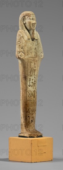 Hornakht's Shabti, back pillars, hieroglyphs, wigs, beards facial hair, Hornakht ca. 525–530 BC, funerary figurine mummy-shaped shabti, grave good, standing on a base, back supported by a pillar/pilaster. Arms crossed on the chest, right folded over the left. In the right hand: a long-bladed hoe and two strings from which hangs a semicircular basket on the back slightly above the elbow; in the left hand: a whip. Hands and tools are engraved. On the head: a long, three-part smooth wig. Long straight beard of even width. Face with coarse features, roughly defined eyes, wide nose, prominent mouth with a slight smile. Right ear significantly larger than the left. Inscription carved in a single column on the front, extending from chin to feet; content unclear but names the deceased Hornakht and his mother. Date: 664–30 BC Late Period–Ptolemaic Period, Egypt. Type: shabti. Dimensions: height 11.6 cm, width 3.1 cm., A small standing figurine with a human face and headdress, shown from a sligh...