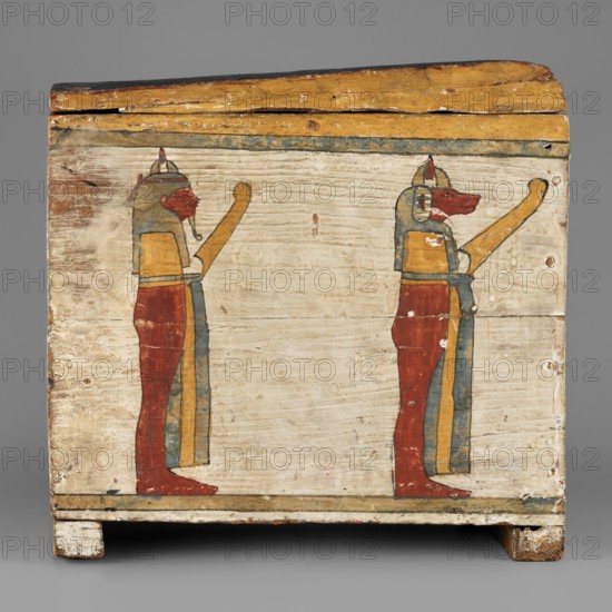 Canopic box, Sons of Horus, falcons, jackals, Isis, Nephthys; grave goods, Egyptian deities; ca. 747–656 BC, Third Intermediate Period, 25th Dynasty, Egypt; canopic box, height 28.5 cm, width 22 cm, depth 29 cm, A rectangular wooden box with a hinged lid sits against a gray background, its painted surface showing a pale off-white ground with a horizontal decorative band near the top in muted blue and yellow tones and a thin darker line near the base; on the front panel are a pair of standing figures facing right, each rendered with red lower garments, yellow upper torsos and arms, blue headdresses and vertical blue-and-yellow draped elements, and red animal-like heads with elongated snouts; the wood grain and aged paint show areas of wear, flaking, and small chips revealing natural wood and darker underlayers, and the box rests on short rectangular feet that show scuffs and discoloration in brown and white.