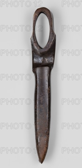 Axe head, unknown Phoenician workshop; Phoenicia Lebanon; 18th–17th century BC. Militaria — edged weapon: axe head with a narrow, elongated blade and a socket for the handle set closer to the butt. Both sides of the socket opening are decorated with a convex shaft in the form of an arch. Weight 610 g., A single elongated metal object photographed against a light gray background, showing a dark brown to nearly black patina with areas of lighter brown rust and wear; the top has an oval opening surrounded by thicker metal, the shaft narrows below that and tapers to a pointed end, with surface irregularities and pitting visible along its length and subtle shine on smoother sections.