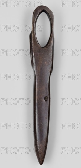 Axe head, unknown Phoenician workshop; Phoenicia Lebanon; 18th–17th century BC. Axe head with a narrow, elongated blade and a stock for the handle closer to the butt. A convex shaft in the form of an arch decorates both sides of the stock opening. Militaria, edged weapon; weapon; weight 610 g., A dark brown elongated metal tool with an oval opening at the top and a tapered pointed end, showing a worn surface with patches of lighter brown and small areas of rust; it is photographed against a light gray background.