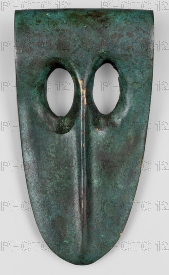 Axe head, unknown Phoenician workshop; militaria, edged weapon; strongly elongated, oval-tipped blade with a thickening along the center; at the opposite end a vertical socket for the base with two elongated oval windows cut into the blade. Phoenicia Lebanon, 20th–17th century BC. Weapon. Height 5.5 cm, width 10.5 cm, depth 2 cm, weight 218 g., An elongated, shield-shaped object with a greenish-blue patina and areas of darker and lighter green, showing mottled brown and black spots, featuring two rounded eye-shaped openings near the top and a raised central ridge running down the middle, with small patches of worn metallic color visible along the ridge and edges.
