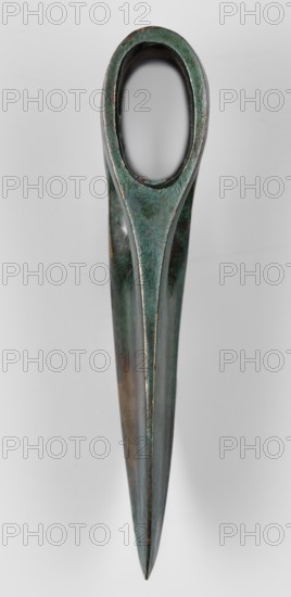 Axe head, unknown Phoenician workshop; militaria, edged weapon; strongly elongated, oval-tipped blade with a thickening along the center. At the opposite end is a vertical socket for the base, in front of which are two elongated oval windows cut into the blade. Phoenicia Lebanon, 20th–17th century BC; height 5.5 cm, width 10.5 cm, depth 2 cm; weight 218 g., A single elongated metal object with a rounded oval opening at the top tapering to a pointed end, showing a textured surface with areas of greenish patina, darker green, brownish spots, and muted metallic gray, photographed against a light gray background.