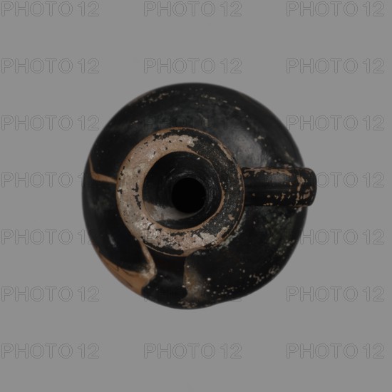 Aryballus lekythos, Attica Greece; Kerch finding. Late 5th–early 4th century BC. Aryballus lekythos with a ring-shaped foot, a bulging belly, and a high neck widening towards the spout. A rounded vice is attached to the body and the middle of the neck. The vessel is covered with black finish. On the surface of the belly, the head of a woman in a cap turned to the right. Red-figure/black-figure pottery attributes: lekythos aryballus lekythos. Vessel; height 7 cm, width 9 cm, depth 5 cm., A round object viewed from above with a central circular opening and a handle extending to one side, showing worn and chipped surfaces with areas of exposed material; the dominant color is black with visible patches of beige and gray, and small speckles of lighter tones across the surface.
