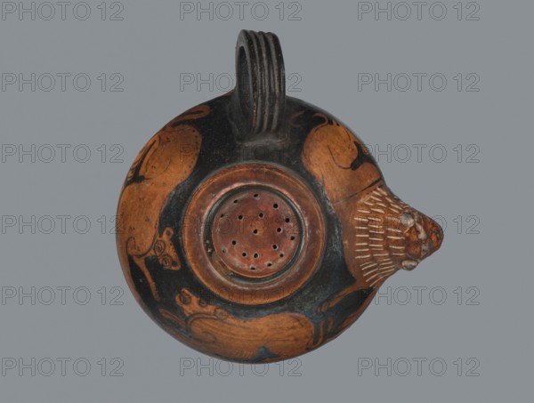 Red-figure guttus, Greece, Attica, 5th century BC c. 425–375 BC. Vessel guttus, height 5.7 cm, width 9.8 cm. Shallow, low cylindrical neck with rim turned outward; rim formed by the plastically modeled muzzle of a lion. Shoulders decorated with a lion and panthers; animals semi-reclining with curled tails, facing left; formed mane and muzzle. Instead of a spout, a strainer is visible at the top; 3 cm wide. Reserved area between the main decorative band and the strainer. Technology: visible turning marks. Varnish matte, dense, good quality; clay purified with a small amount of mica. Surface contaminated. Clay color 2.5 YR 5/6 red; varnish color 10 YR 2/1 black., A round ceramic vessel shown from above with a looped handle at the top and a protruding spout shaped like an animal head on the right, decorated with painted animal figures and patterns. The surface shows a mix of reddish-brown and black colors, with lighter tan highlights on the animal head and decorative lines. The central re...
