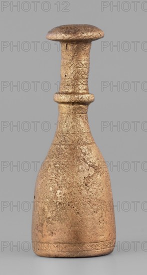 Bottle-type idol base, unknown Luristan workshop. Bottle-shaped base with a long, narrow neck terminating in an upturned, slightly convex spout. Halfway up, a convex cylinder with an engraved zigzag line encircles the neck. Engraved decoration of horizontal bands of line motifs—diagonal crosses, chevrons, hatched triangles—on the central part of the neck and at the lower edge of the base. The base is open at the bottom and hollow inside; a sleeve-shaped hole passes through the neck to allow mounting of a pin with an idol figurine. Geometric ornaments; bottle base. Iron Age II, 8th–7th century BC. Luristan, Iran. Base; height 7.5 cm, width 2.8 cm., A single sculpted object resembling a bottle or small vessel stands upright against a plain grey background; the object has a wide rounded base that tapers into a narrower neck topped by a flattened, mushroom-like rim and a pronounced collar around the midneck. The surface appears rough and textured with visible pitting and irregularities, an...