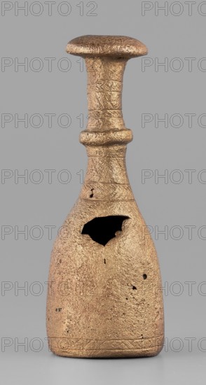Bottle-type idol base, unknown Luristan workshop. A bottle-shaped base with a long, narrow neck terminating in an upturned, slightly convex spout. Halfway up, a convex cylinder with an engraved zigzag line encircles the neck. Engraved decoration of horizontal bands of line motifs—diagonal crosses, chevrons, hatched triangles—appears on the central part of the neck and at the lower edge of the base. The base has no bottom and is hollow inside; a sleeve-shaped hole passes through the neck to allow mounting of a pin with an idol figurine. Iron Age II, 8th–7th century BC. Luristan, Iran. Base; height 7.5 cm, width 2.8 cm., A single metallic object with a textured surface stands against a plain gray background, showing a warm bronze color with areas of darker and lighter bronze tones; the object has a rounded, flattened top connected to a narrow neck with a banded ridge, widening into a bell-shaped body that tapers to a flat base, and the surface exhibits a few irregular pits and a large ja...