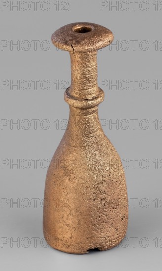 Bottle-type idol base, unknown Luristan workshop. A bottle-shaped base with a long, narrow neck terminating in an upturned, slightly convex spout. Halfway up, a convex cylinder with an engraved zigzag line encircles the neck. Engraved decoration of horizontal bands of line motifs—diagonal crosses, chevrons, hatched triangles—appears on the central part of the neck and at the lower edge of the base. The base is open no bottom and hollow inside, with a sleeve-shaped hole through the neck to allow mounting of a pin with an idol figurine. Geometric ornaments; bottle base. Iron Age II, 8th–7th century BC. Luristan, Iran. Height 7.5 cm; width 2.8 cm., A tall, narrow object with a textured surface in shades of brown and bronze, featuring a flared top with a central hollow, a slender neck with a raised ring around it, and a broader rounded base that shows surface irregularities and roughness against a plain gray background.