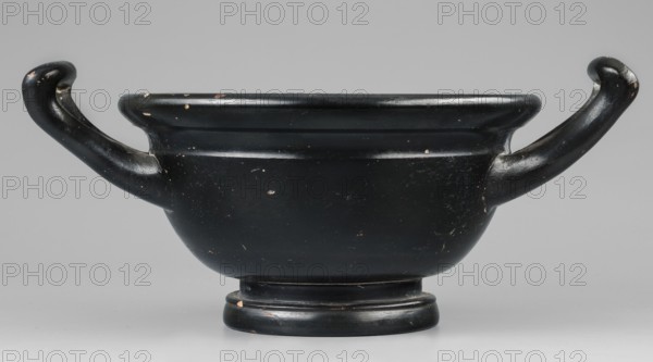 Black-finned skyphos, early 4th century BC, Attica. Thick-walled skyphos with two handles, deep bowl on a low profiled foot. Interior: stamped ornament with six palmettes; inside the circle two concentric circles with ovules between them; surrounded by a band of ovules between two engraved lines. Outer part of the foot decorated with concentric circular elements. Technology: evenly distributed varnish, matte, visible turning marks; admixtures: mica, pyroxene. Clay color 2.5 YR 6/6 light red. Varnish color 10 YR 2/1 black. Vessel skyphos. Dimensions: height 10.4 cm; width 23.5 cm; depth 16.6 cm; weight 0.456 kg; foot diameter 8.2 cm., A shallow, wide bowl with a rounded base and a short pedestal stands centered in the image, finished in a glossy black with small areas of surface wear revealing a lighter color beneath; the bowl has a broad rim and a pair of sweeping, curved handles extending outward and upward from opposite sides, each handle sharing the same black finish and exhibiting ...
