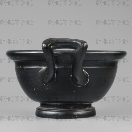 Black-finned skyphos, early 4th century BC, Attica. Thick-walled skyphos with two handles, deep bowl on a low profiled foot. Interior decorated with a stamped ornament of six palmettes; inside the circle are two concentric circles with ovules between them, surrounded by a band of ovules between two engraved lines. Outer part of the foot decorated with concentric circular elements. Technology: evenly distributed matte varnish, visible turning marks; admixtures: mica, pyroxene. Clay color 2.5 YR 6/6 light red. Varnish color 10 YR 2/1 black. Vessel skyphos, height 10.4 cm, width 23.5 cm, depth 16.6 cm, weight 0.456 kg, foot diameter 8.2 cm., A black bowl-like vessel with a rounded body and a flared rim, shown from the front and centered against a smooth gray background; the vessel rests on a short circular foot and has a pair of stout looped handles attached near the rim, with visible wear and small lighter-colored specks on the black surface.