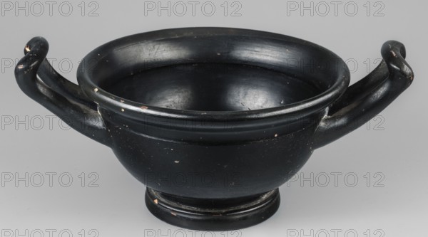 Black-finned skyphos, early 4th century BC, Attica; thick-walled variety with two handles, deep bowl on a low profiled foot. Interior: stamped ornament with six palmettes; inside the circle two concentric circles with ovules between them; surrounded by a band of ovules between two engraved lines. Outer part of the vessel's foot decorated with concentric circular elements. Technology: varnish evenly distributed, matte; visible turning marks. Admixtures: mica, pyroxene. Clay color 2.5 YR 6/6 light red. Varnish color 10 YR 2/1 black. Vessel skyphos: height 10.4 cm, width 23.5 cm, depth 16.6 cm, weight 0.456 kg, foot diameter 8.2 cm., A ceramic bowl with a rounded body and a flared rim shown against a neutral background, finished in a glossy black surface with a few small worn spots revealing a lighter color beneath; the bowl has a pedestal base and a pair of curved handles that extend outward and curl at the ends, also black with some scuffed areas, and the interior is smooth and black wi...