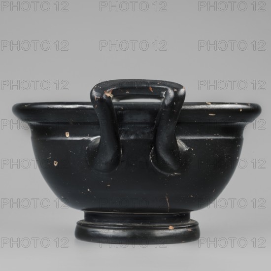 Black-finned skyphos, Greek ceramics, early 4th century BC, Attica, thick-walled cup skyphos with deep bowl on a low profiled foot. Interior stamped ornament with six palmettes; inside the circle two concentric circles with ovules between them; surrounded by a band of ovules between two engraved lines. Outer part of the foot decorated with concentric circular elements. Varnish evenly distributed, matte, visible turning marks; admixtures: mica pyroxene. Clay color 2.5 YR 6/6 light red; varnish color 10 YR 2/1 black. Vessel: height 10.4 cm, width 23.5 cm, depth 16.6 cm, weight 0.456 kg, foot diameter 8.2 cm., A black glazed ceramic bowl with a rounded body and a flared rim, shown from the front on a neutral gray background; the bowl has a pair of looped handles that arc from the rim to the shoulder, a raised circular foot, and several small chips and worn spots revealing a lighter color beneath; colors visible include black, gray, and small areas of a light beige where the glaze is worn.