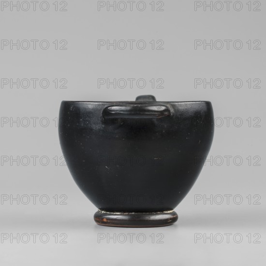 Black-finished skyphos, Greek ceramics, Type B. Thin-walled skyphos with a deep bowl and annular foot. One handle mounted horizontally, the other vertically. Entirely covered with a high-quality, glossy black varnish; only the bottom remains clay-colored with decoration of concentric circles. Technology: Visible traces of turning. Small chips on the surface. Clay color 2.5 YR 5/6 red. Varnish color 10 YR 2/1 black. Attributed to Attica, 480–450 BC. Vessel skyphos. Height 7.6 cm; width 14.1 cm; depth 8.8 cm; weight 0.102 kg., A smooth, rounded dark object with a glossy black surface sits centered against a light gray background, showing a slightly bulbous body tapering to a small circular base with hints of brown and worn areas; a short horizontal handle-like protrusion is visible near the top of the body, and the surface exhibits small scuffs and subtle reflections in shades of black and very dark brown.