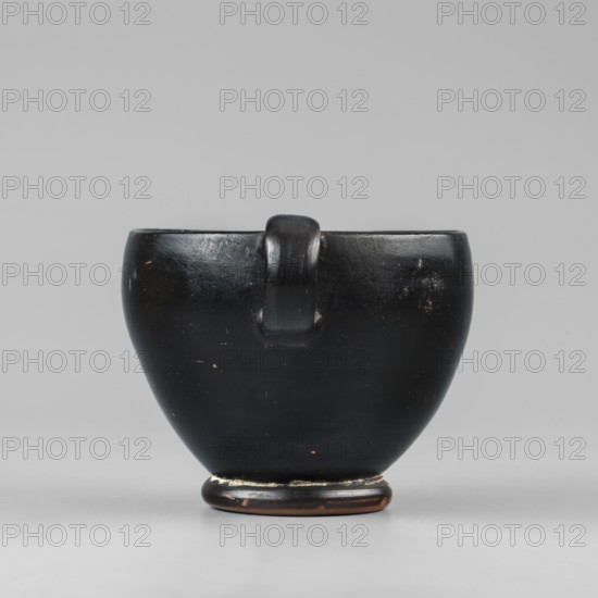 Black-finished skyphos, Type B. Thin-walled skyphos with a deep bowl and annular foot. One handle mounted horizontally, the other vertically. Entirely covered with a high-quality, glossy black varnish; only the bottom remains clay-colored with decoration of concentric circles. Technology: Visible traces of turning. Small chips on the surface. Clay color 2.5 YR 5/6 red. Varnish color 10 YR 2/1 black. Attributed to Attica, 480–450 BC. Vessel skyphos. Height 7.6 cm; width 14.1 cm; depth 8.8 cm; weight 0.102 kg., A smooth, rounded object with a glossy black surface and a small rounded handle at the top center sits on a low circular base with worn areas revealing brown beneath; the background and surface beneath the object are light gray, and the object shows a few small scuffs and lighter spots on its black finish.