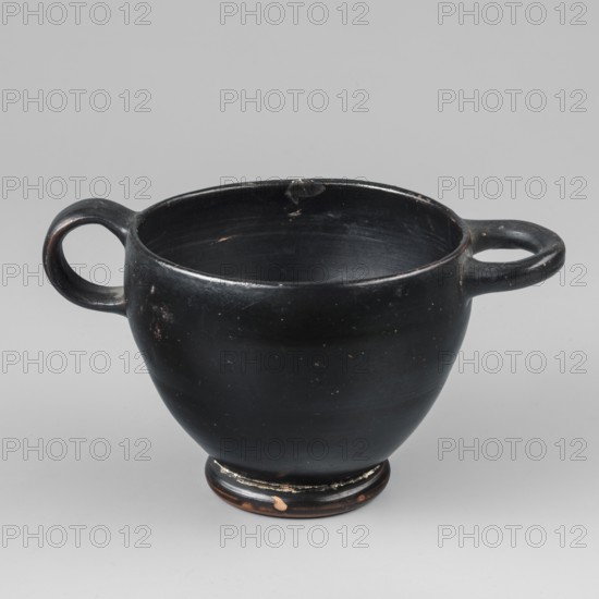 Black-finished skyphos, Type B. Thin-walled skyphos with a deep bowl and annular foot. One handle mounted horizontally, the other vertically. The object is entirely covered with a high-quality, glossy black varnish; only the bottom remains clay-colored, decorated with concentric circles. Technology: visible traces of turning; small chips on the surface. Clay color: 2.5 YR 5/6 red. Varnish color: 10 YR 2/1 black. Attributed to Attica, 480–450 BC. Vessel skyphos. Height 7.6 cm; width 14.1 cm; depth 8.8 cm; weight 0.102 kg., A black ceramic cup with a rounded body and a low foot, featuring a pair of horizontal loop handles on opposite sides, sits against a light gray background; the cup shows small worn spots and areas where the black finish is chipped to reveal a brownish color underneath, and the interior and rim display subtle scuffs and lighter marks.