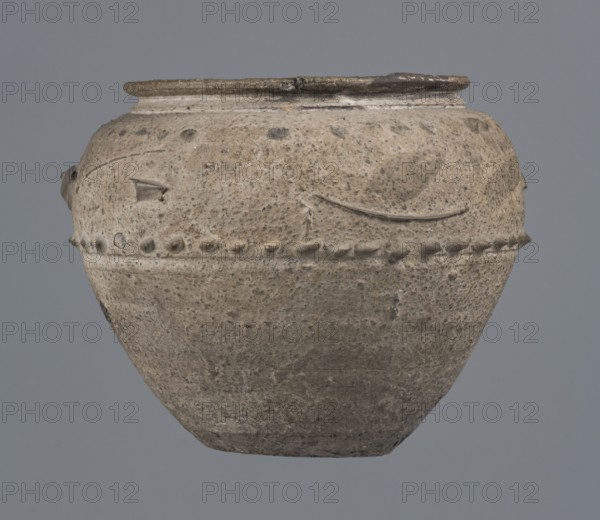 Vise-free cup, unknown Rhine workshop, Goluchów Greater Poland Voivodeship. Hard-fired, granular, light gray clay 7.5YR N7/; gray matte glaze 10YR 5/18 covering the entire surface, intentionally rough sandblasted. Upper body with en barbotine decoration of stylized lanceolate leaves between two stripes of dots. Whole vessel, 2nd half of the 1st century, Cologne, height 8.0 cm, width 7.0 cm., A weathered rounded pot with a wide flattened rim sits against a plain background; the surface shows a coarse, pitted texture with a band of raised bead-like bumps encircling its midsection and some shallow incised lines and chips near the upper area; the pot displays mottled tones of beige, light brown, gray, and darker brown speckles, with subtle variations in those colors across its body.