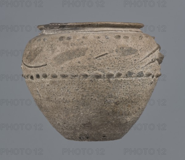 Vise-free cup, unknown Rhine workshop, Goluchów, 2nd half of the 1st century, possibly Cologne. Hard-fired, granular, light gray clay 7.5YR N7/; gray matte glaze 10YR 5/18 covering entire surface, intentionally rough sandblasted. En barbotine decoration on upper body: stylized lanceolate leaves between two stripes of dots. Whole vessel; height 8.0 cm, width 7.0 cm., A rounded, textured ceramic vessel with a narrow rim and a bulbous body sits against a plain background; the vessel surface shows a rough, speckled texture and an overall beige to gray color with darker gray spots and patches, a few raised decorative elements form a horizontal band around the upper third, and there are several subtle incised lines and small protrusions near the rim, with areas of slightly darker gray and brownish discoloration.