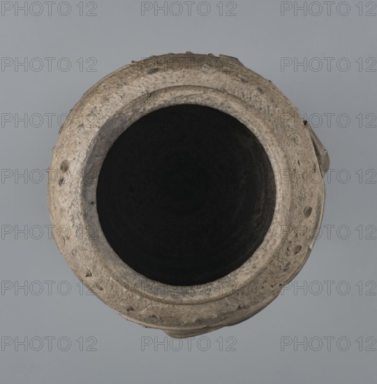 Vise-free cup, unknown Rhine workshop, Goluchów Greater Poland Voivodeship. Hard-fired, granular, light gray clay 7.5YR N7/; gray matte glaze 10YR 5/18 covering entire surface, intentionally rough sandblasted. Upper body with en barbotine decoration of stylized lanceolate leaves between two stripes of dots. Whole vessel, 2nd half of the 1st century, Cologne, height 8.0 cm, width 7.0 cm., A top-down view of a round clay vessel with a wide dark interior and a rough textured rim showing irregularities and small protrusions, in shades of beige, tan, gray, black, and brown against a muted gray background.