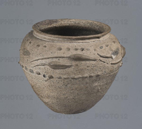 Vise-free cup, unknown Rhine workshop, Goluchów Greater Poland Voivodeship. Hard-fired, granular, light gray clay 7.5YR N7/; gray glaze 10YR 5/18, matte, covers entire surface, intentionally rough sandblasted. On upper body, en barbotine decoration of stylized lanceolate leaves between two stripes of dots. Whole vessel, 2nd half of the 1st century, Cologne, vessel, height 8.0 cm, width 7.0 cm., A rounded clay vessel with a wide open mouth and a flared rim, showing a rough, pitted surface in shades of beige, tan, gray, and brown. The body of the vessel features raised decorative ridges and a row of small rounded protrusions encircling it, along with a few elongated, leaf-like applied elements. The interior appears darker, almost charcoal gray, contrasting with the lighter exterior. The vessel sits against a smooth, muted bluish-gray background.